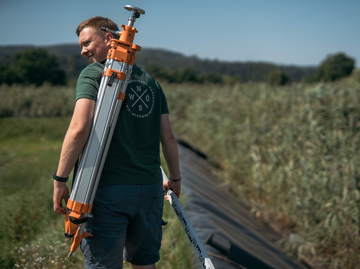 Mann in grünem T-shirt läuft mit einem Werkzeug an einem Schilfbeet entlang