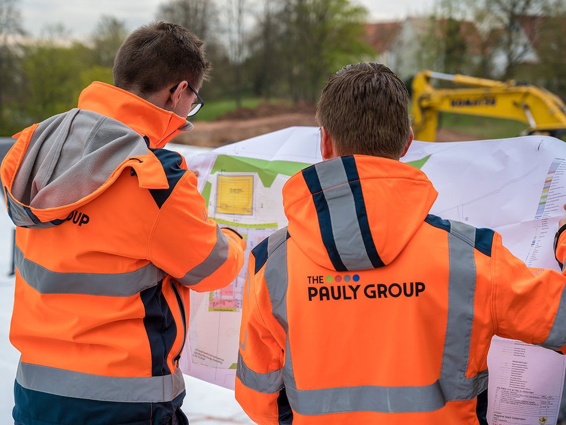 Zwei Mitarbeiter in orangener PAULY GROUP Jacke halten einen Bauplan