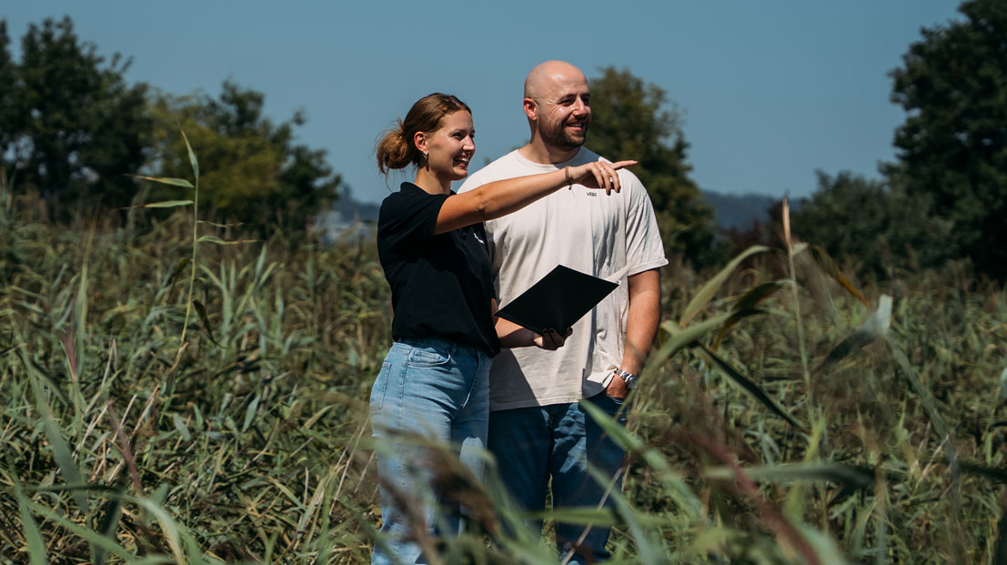 Mitarbeiter auf Schilfbeet mit Mappe in der Hand, Frau zeigt auf etwas